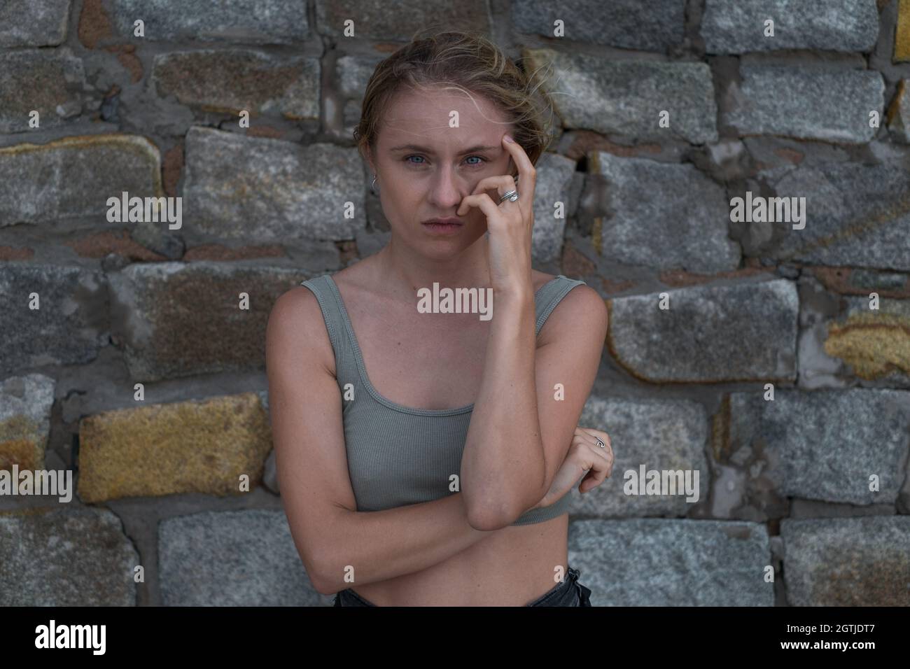 Sad pensive face of young caucasian blonde woman in crop top on the stone background. Young woman crying and holding her hand near the face, sad Stock Photo