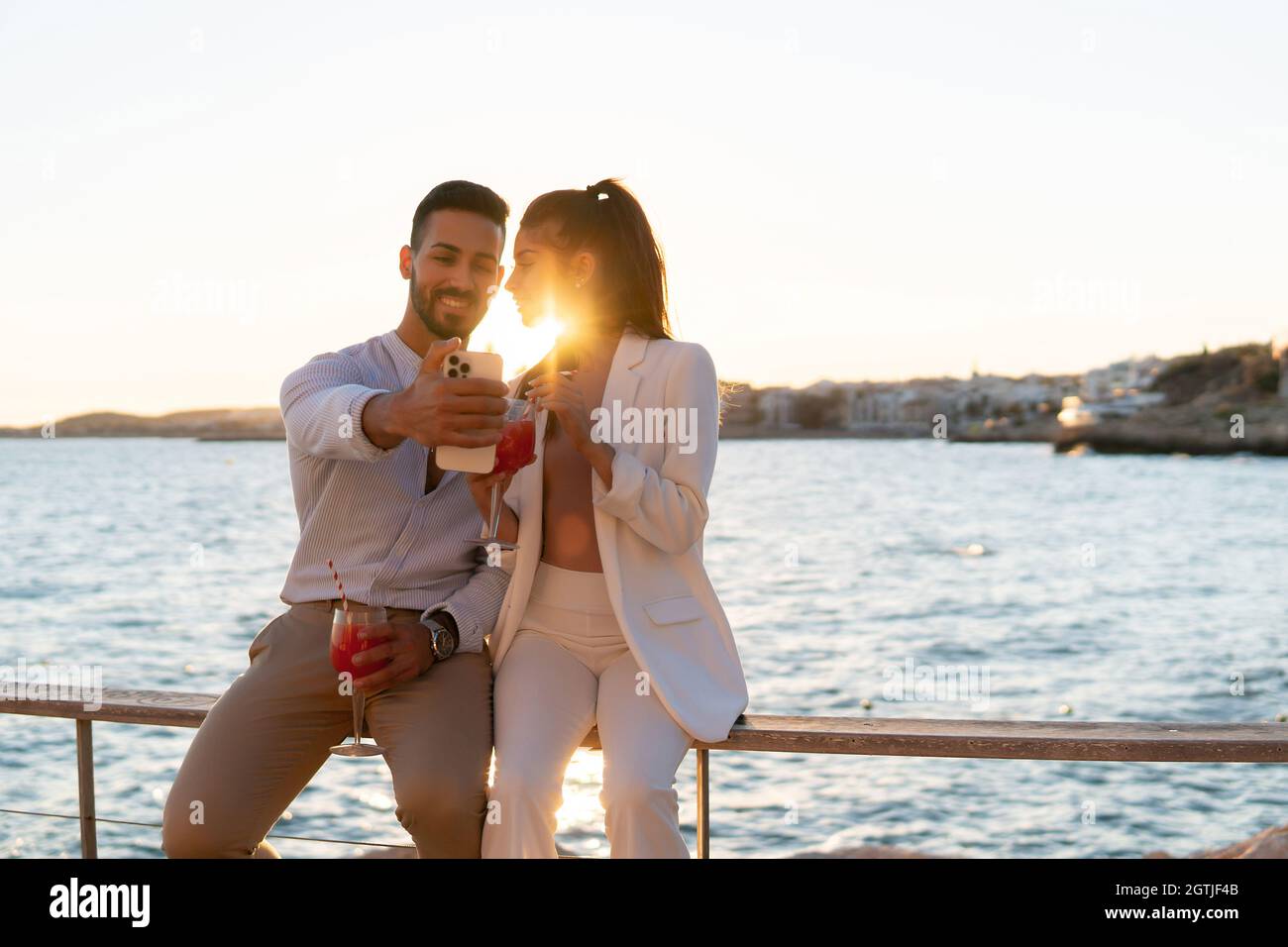 Tender stylish Hispanic couple sitting on embarkment with cocktails and taking selfie on smartphone against sunset sky and sea Stock Photo