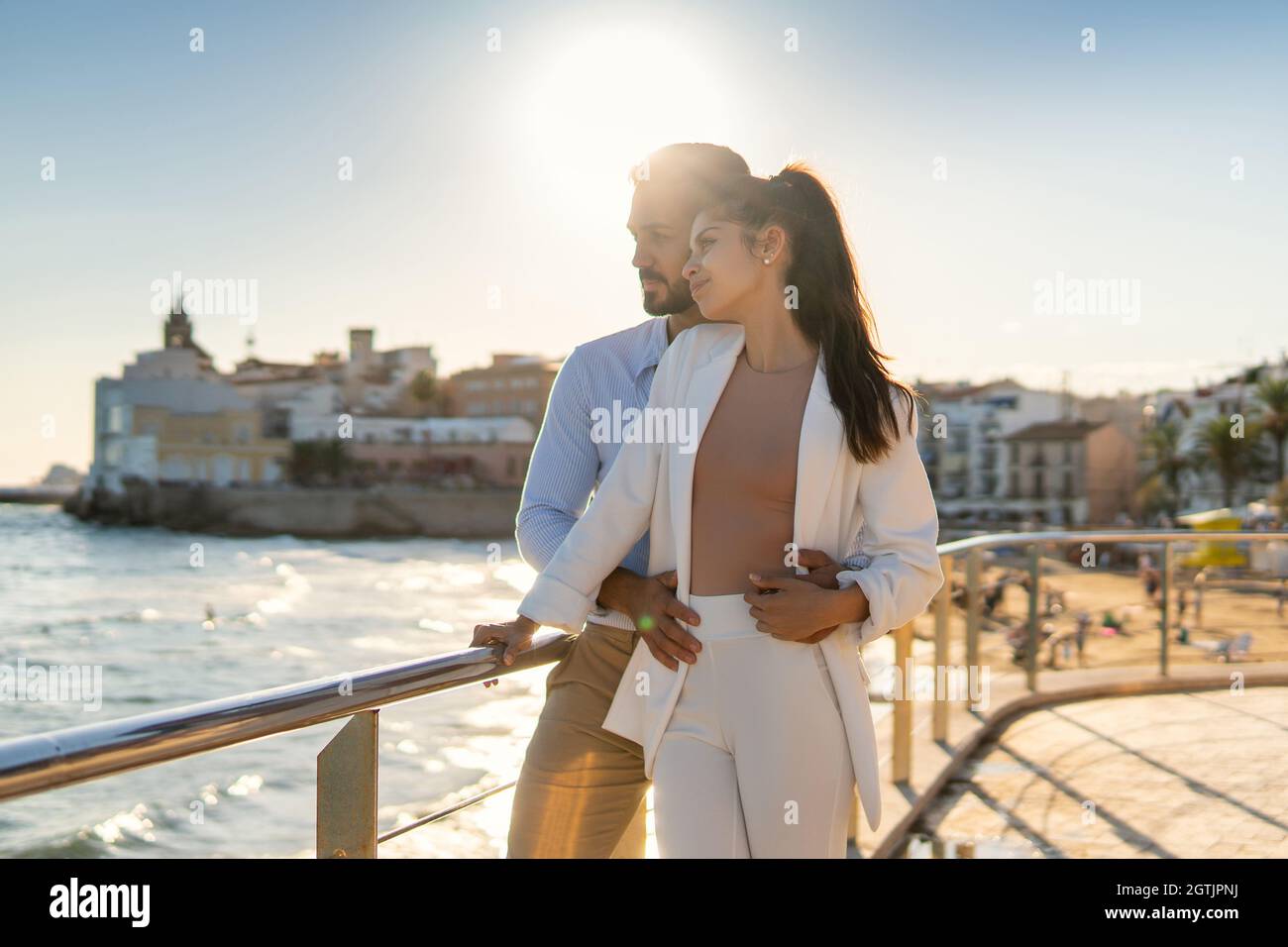 Loving delicate Hispanic couple hugging and standing on promenade near sea while enjoying sunny day in summer and looking away Stock Photo