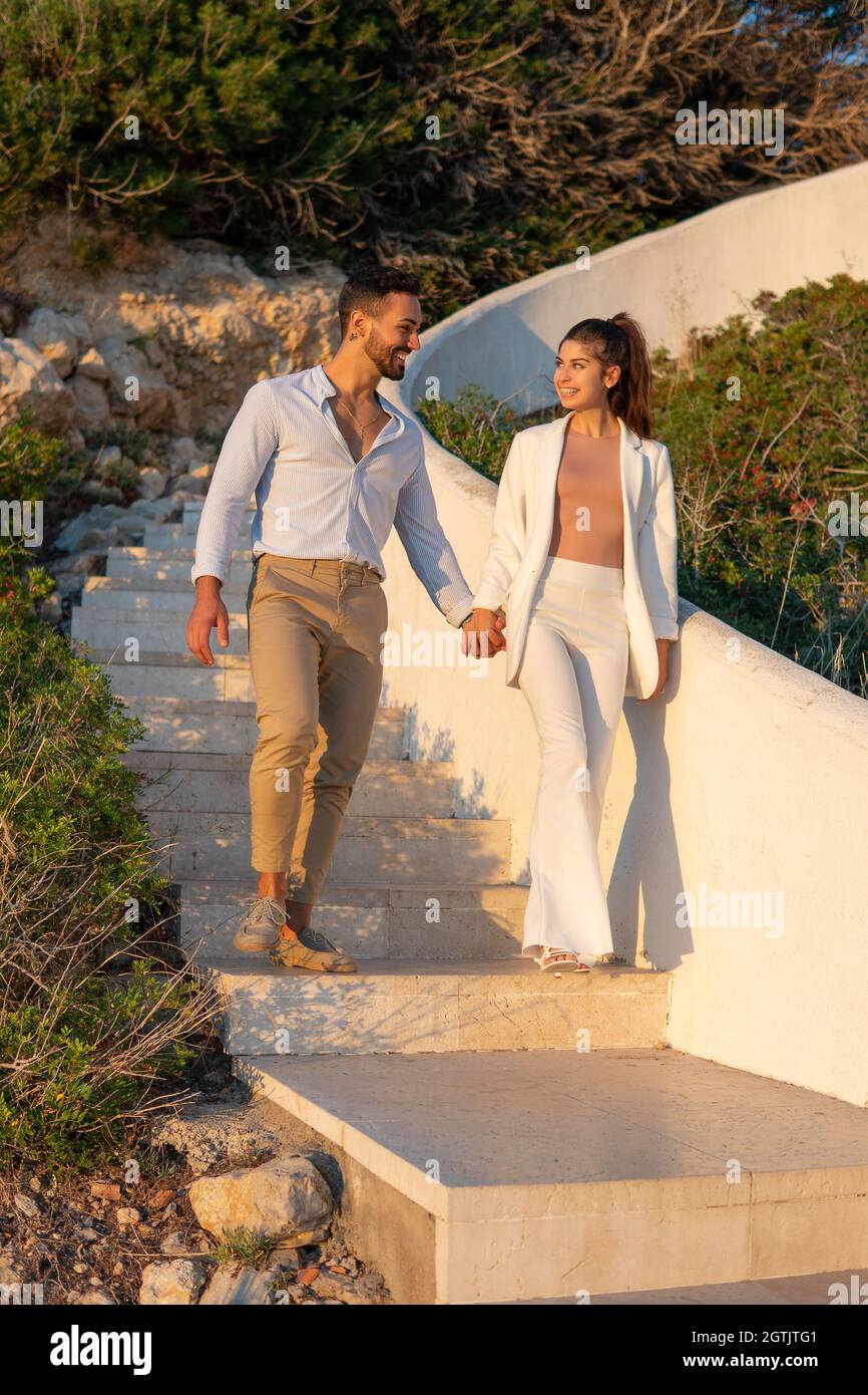 Cheerful loving Hispanic couple holding hands and walking down stone stairs in park while looking at each other and enjoying stroll in summer evening Stock Photo