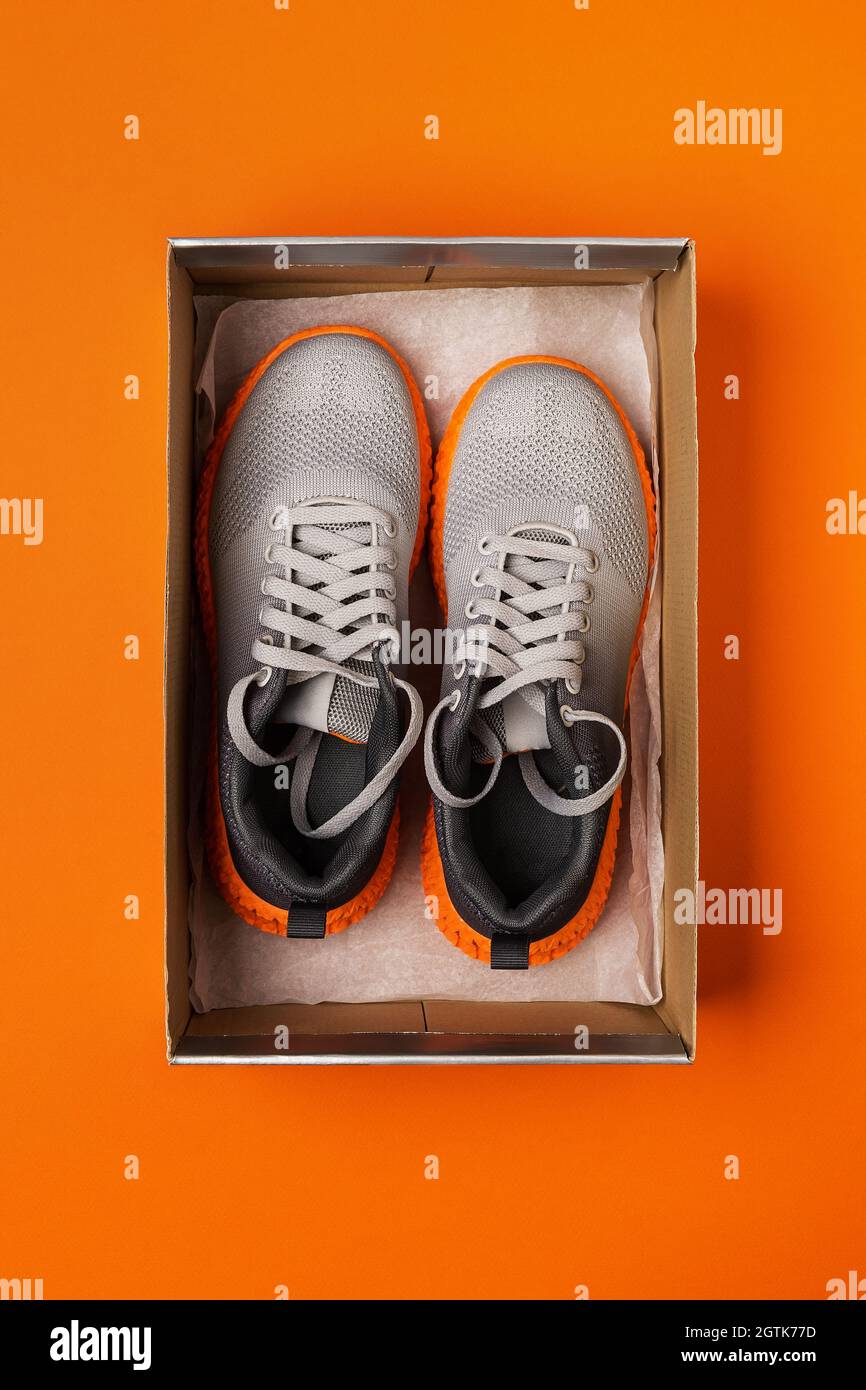 Pair of new gray mesh fabric sneakers in the open box on the bright orange background. Modern textile trainers with grooved orange sole for sport. Stock Photo