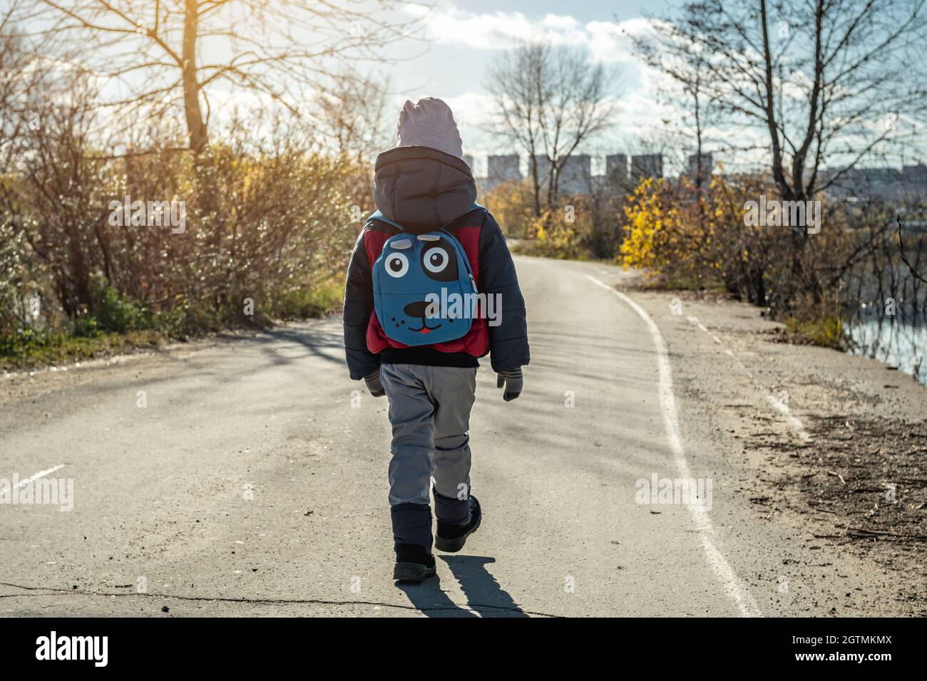 A child boy hiking with a small backpack is walking along the road outside the city on the background of the autumn landscape Stock Photo