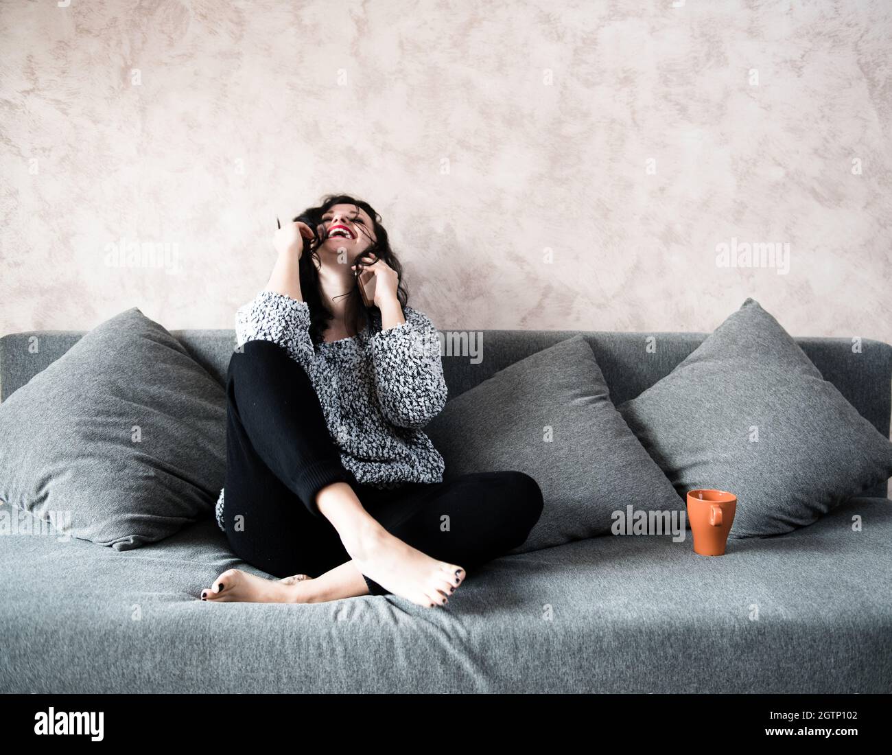 Cheerful Young Woman Answering Smart Phone While Sitting On Sofa At Home Stock Photo