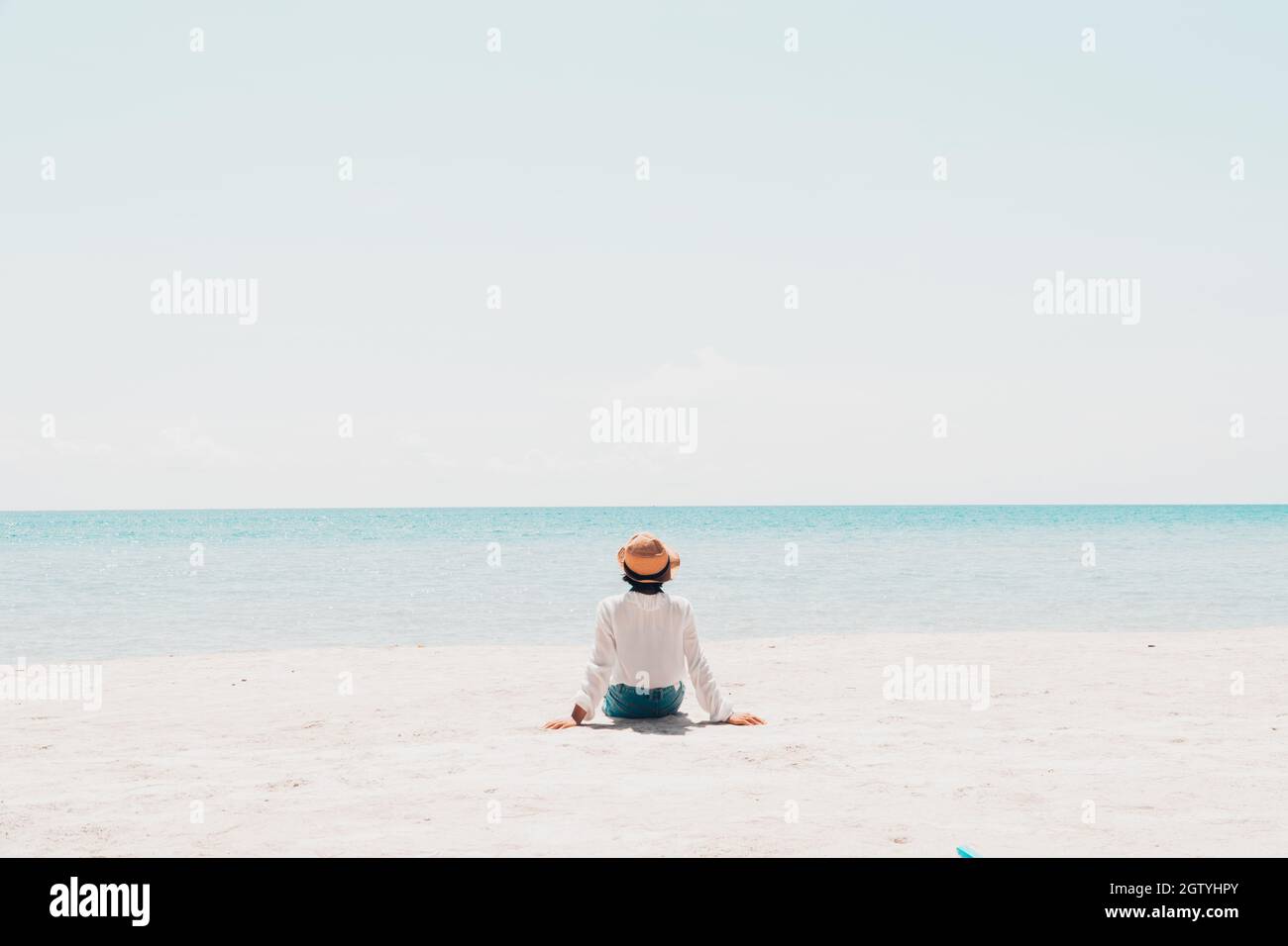 Rear View Of Girl Sitting At Beach Against Clear Sky During Sunny Day Stock Photo