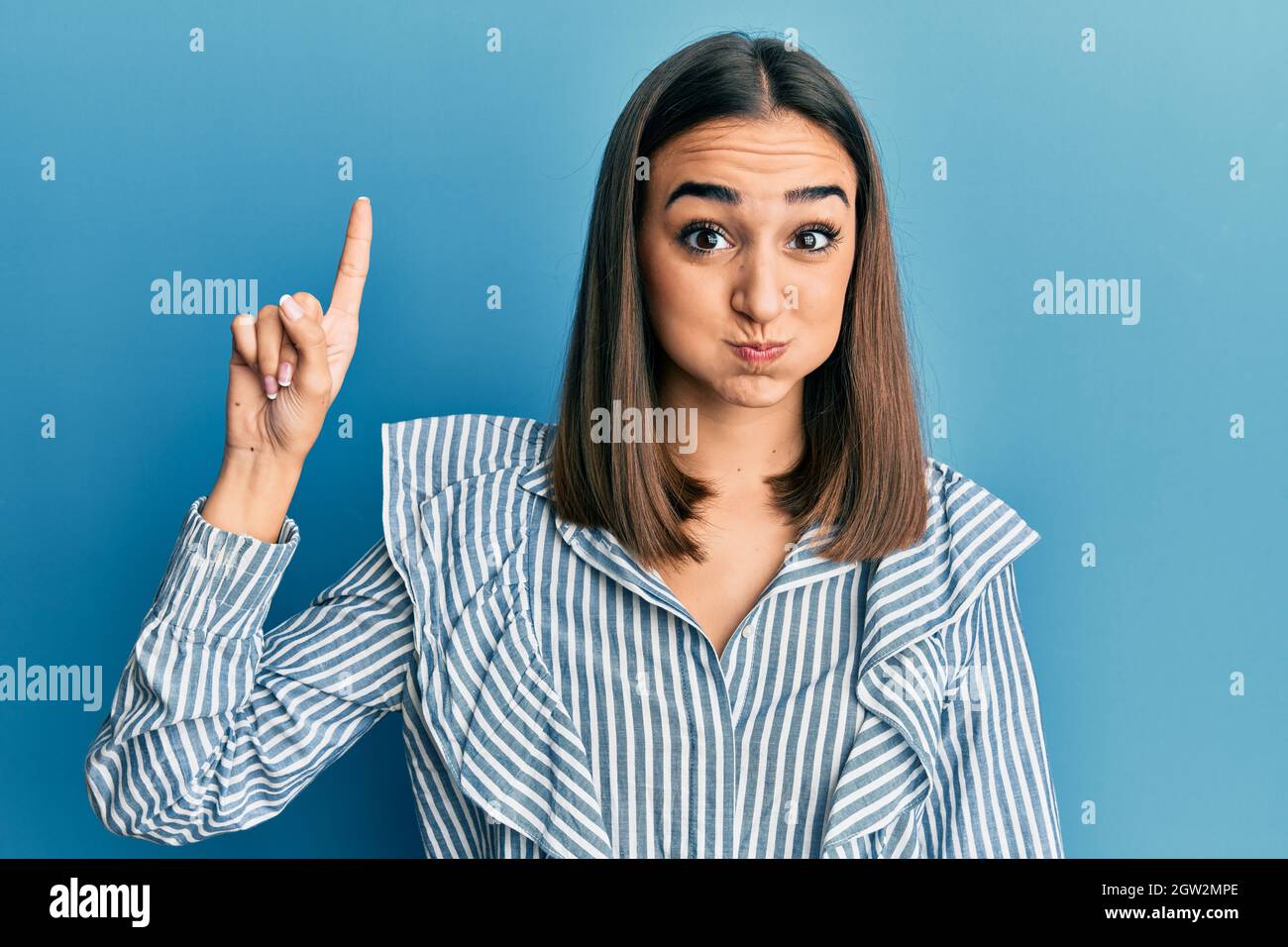 Young brunette student girl pointing up with finger puffing cheeks with funny face. mouth inflated with air, catching air. Stock Photo