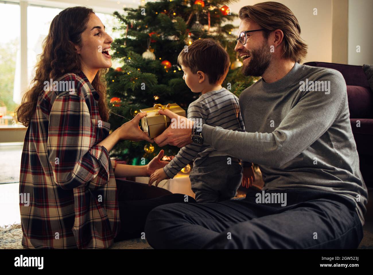 Woman receiving a gift for christmas and new year from her husband. Happy young family sitting by Christmas tree on christmas eve. Stock Photo