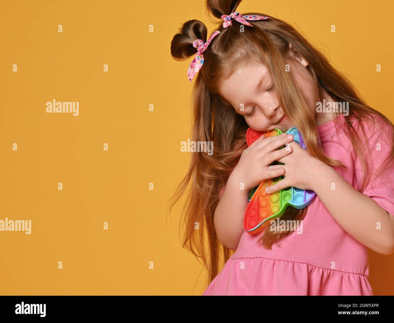 Cute red-haired kid girl in pink shirt hugs new sensory rainbow color butterfly shape toys - pop it Stock Photo