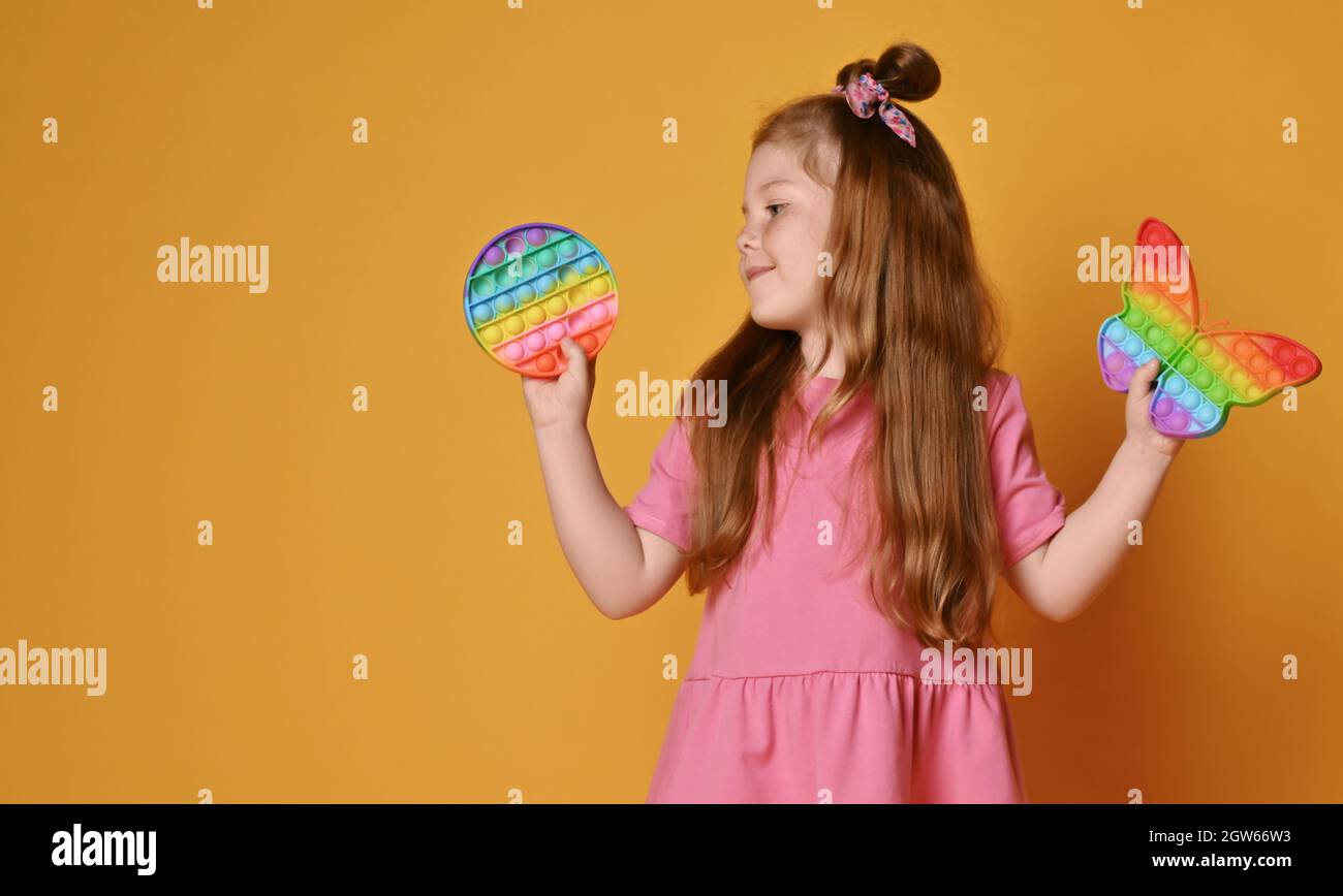 Redhead kid girl in pink clothes holds two sensory rainbow color round and butterfly shape toys - pop it, looking at it Stock Photo