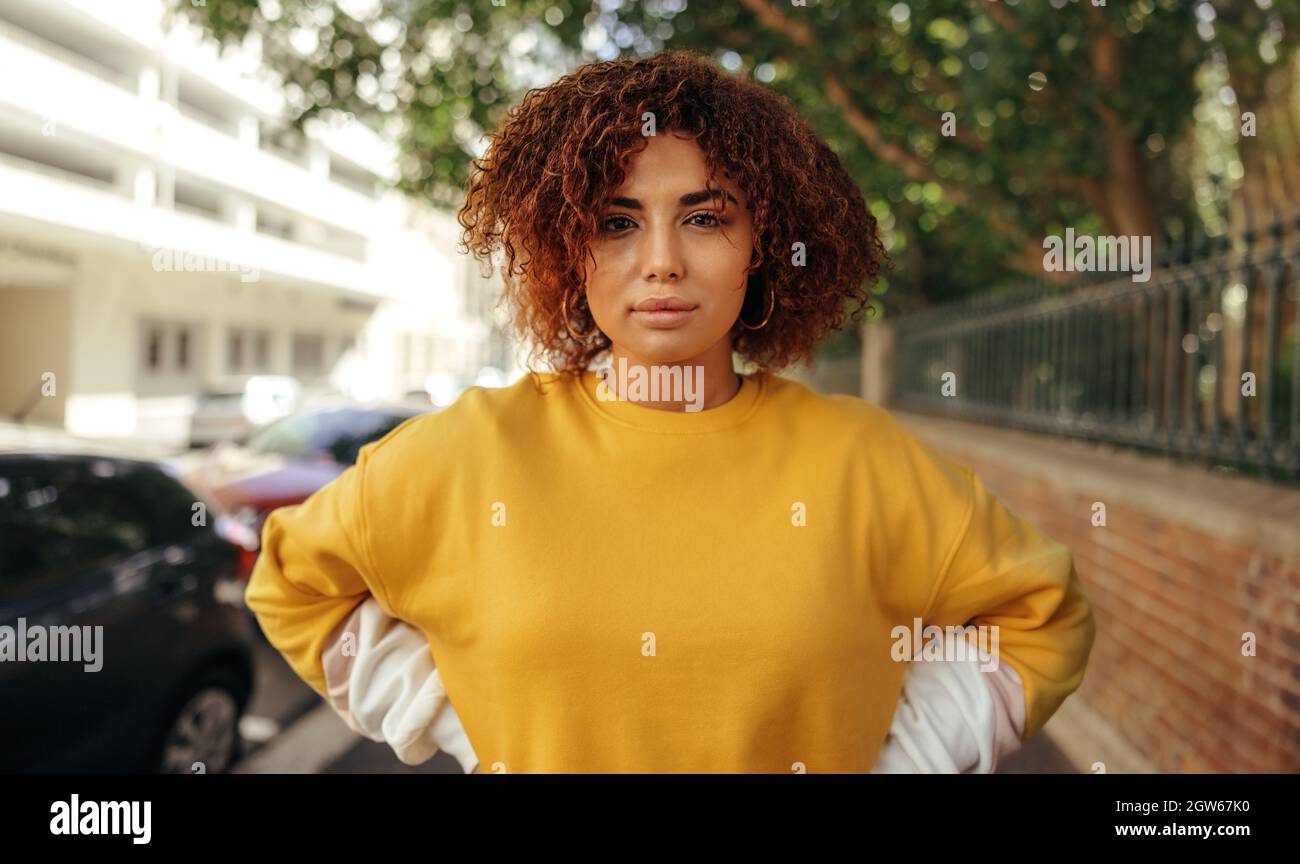 Self-confident teenage girl looking at the camera. Female youngster wearing red hair and a mustard sweatshirt in the city. Young teenager standing on Stock Photo