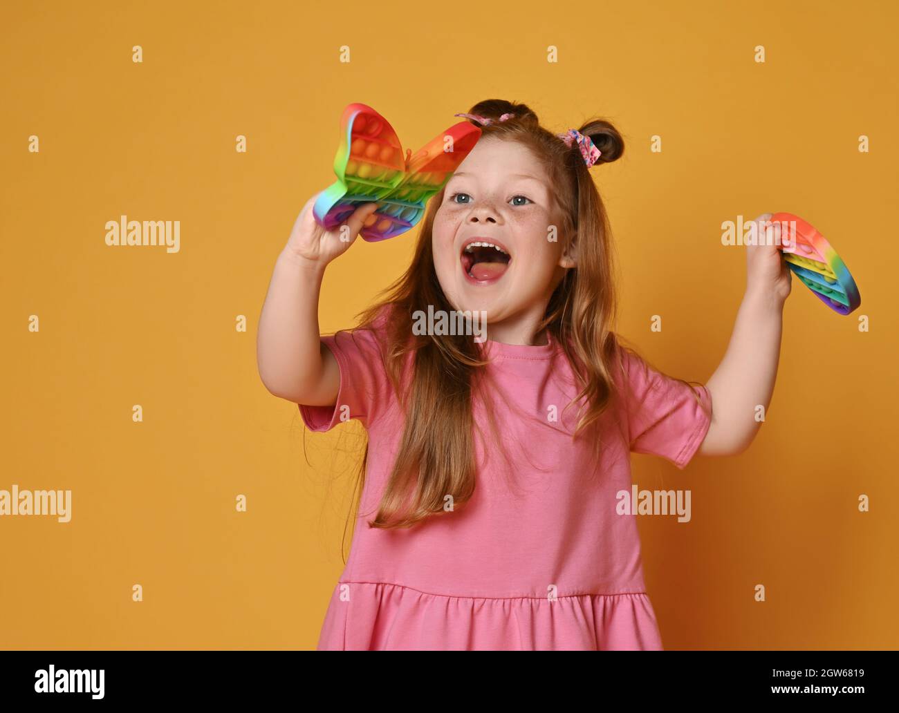 Red-haired kid girl in pink shirt plays with new sensory rainbow color toys - butterfly shape and round pop it Stock Photo
