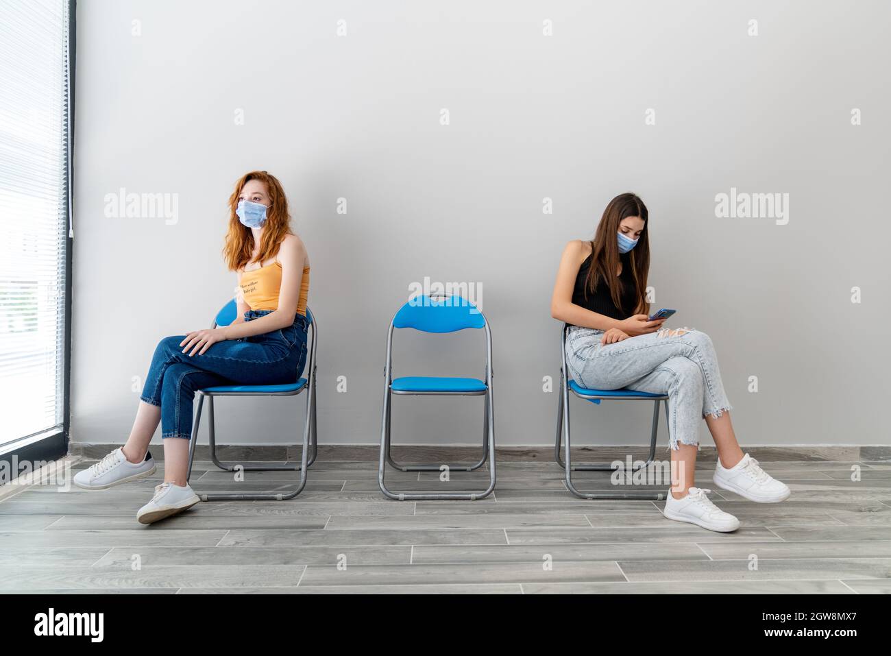 Two young woman with protection mask is sitting in a waiting hall with a distance. Corona virus outbreaking. High quality photo Stock Photo