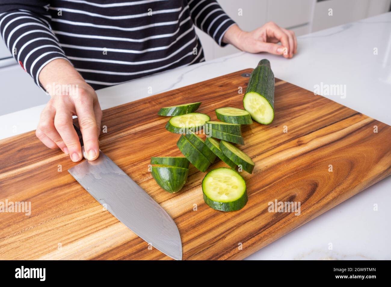 Caucasian Woman Holding Chef Knife On Wooden Cutting Board With Sliced Cucumber In Modern Kitchen Stock Photo