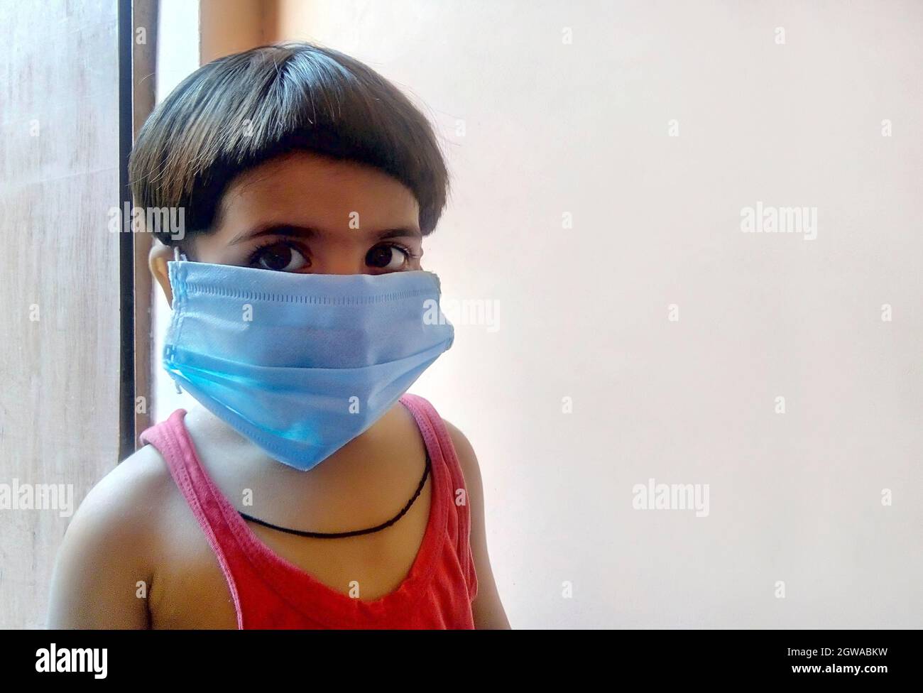 Indian Kid Wearing Protective Face Mask While Standing Against Door At Home Stock Photo