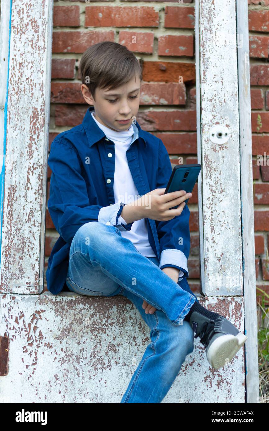 A teenager boy in a blue shirt and jeans with a smartphone in his hands sits on an old door against a brick wall background. Selective focus Stock Photo