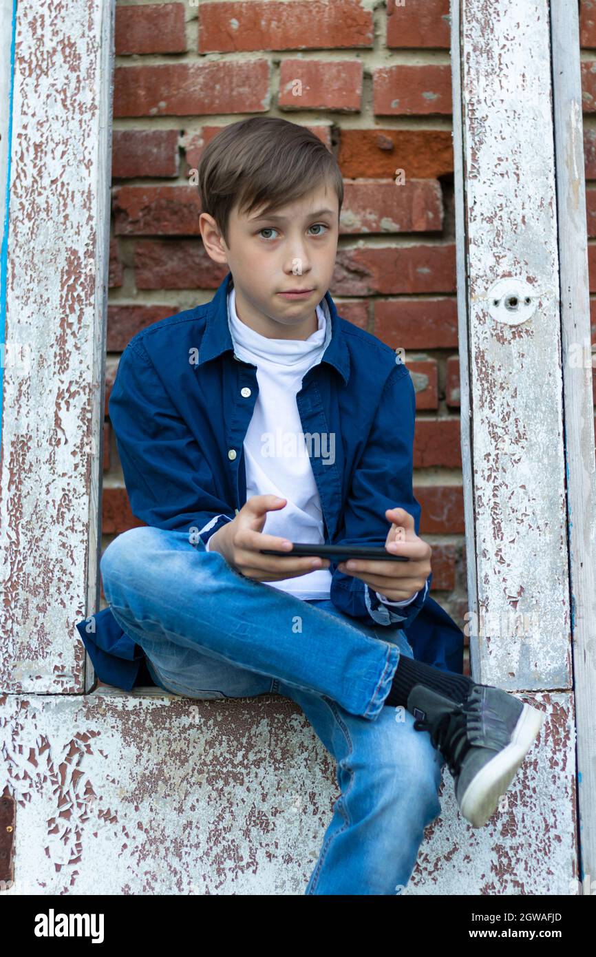 A teenager boy in a blue shirt and jeans with a smartphone in his hands sits on an old door against a brick wall background. Selective focus Stock Photo