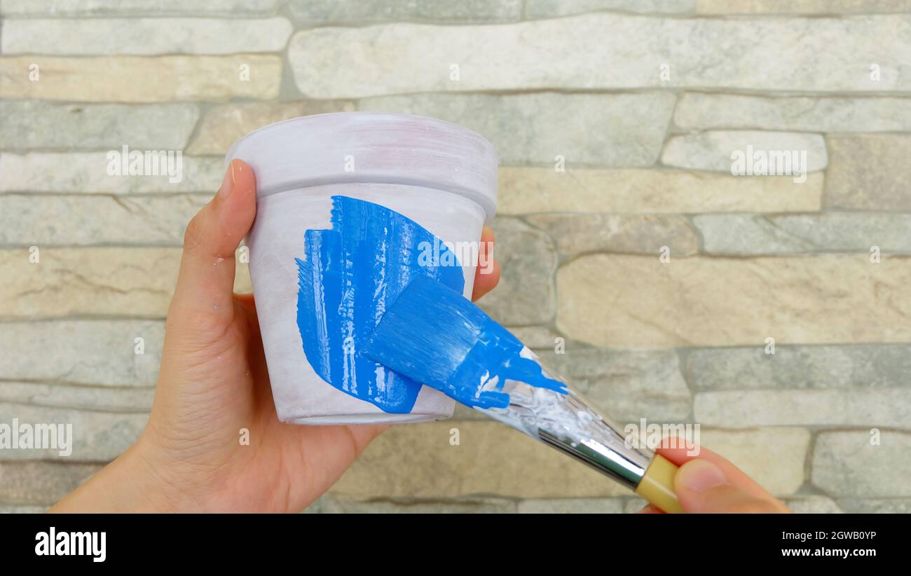 Hand holding a terracotta pot with white base paint, with another hand holding a blue paint brush, coloring the pot with blue paint. Stock Photo