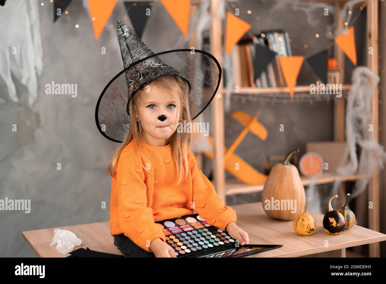 Portrait of a little girl in a Halloween costume sitting at the table with a palette of eyeshadows for makeup in a witch hat. Holiday on October 31 Stock Photo