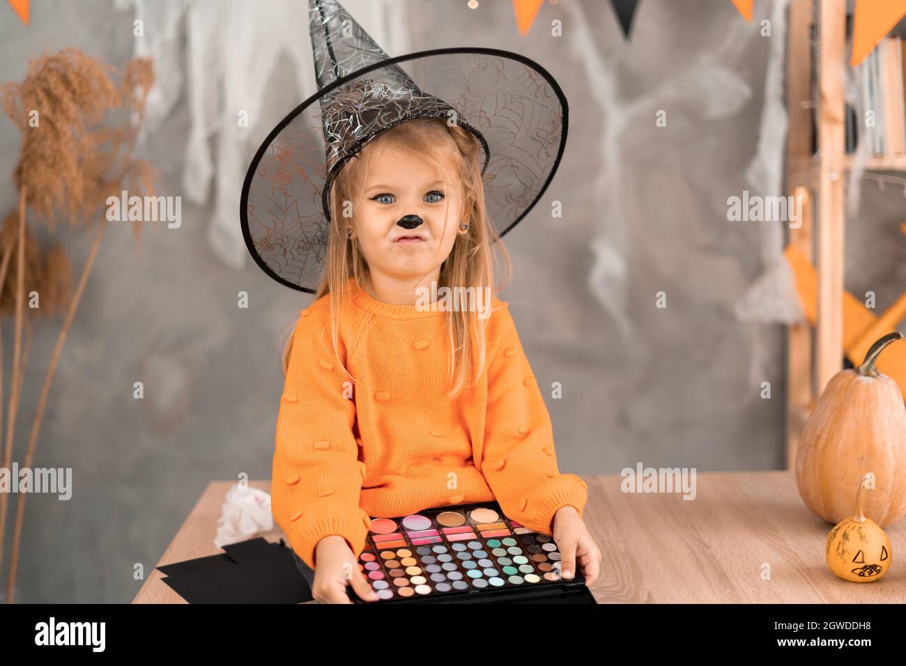 Halloween makeup little girl in the room on the table with a palette of shadows in his hands is preparing for the holiday. a child in a witch hat. Stock Photo