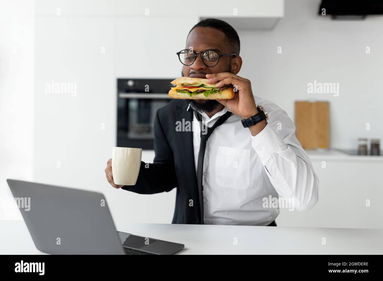Modern Busy Lifestyle. Portrait of African male in eyeglasses having breakfast in kitchen at home, wearing suit and drinking coffee while eating sandw Stock Photo