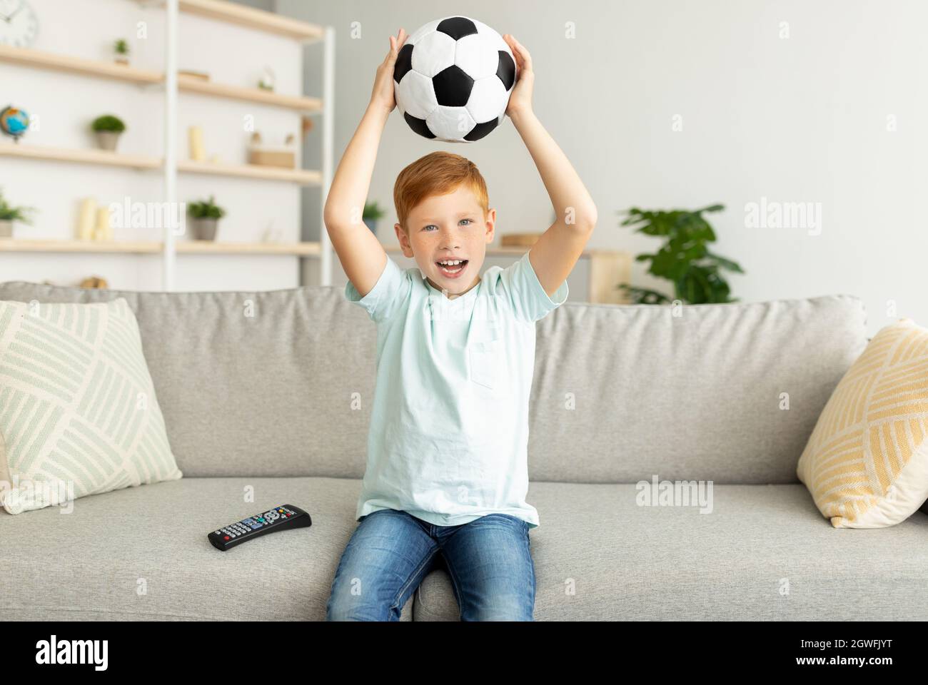 Emotional redhead preteen boy with ball sitting on couch at home, holding ball and watching football game on TV alone, enjoying his soccer team game, Stock Photo