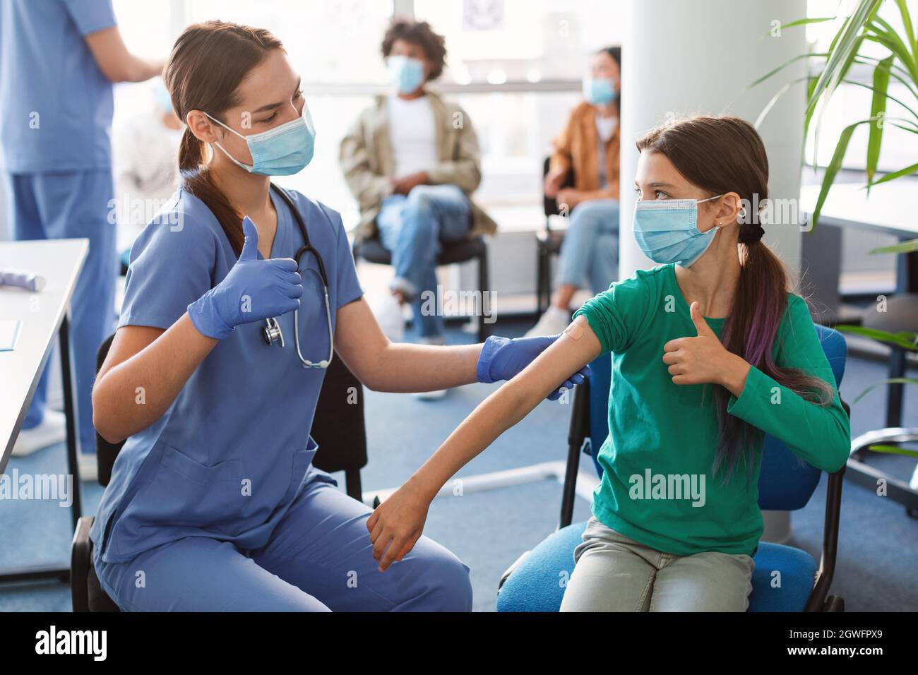 Portrait of adolescent patient in protective face mask showing thumbs up gesture after getting injection at hospital. Doctor applying adhesive bandage Stock Photo