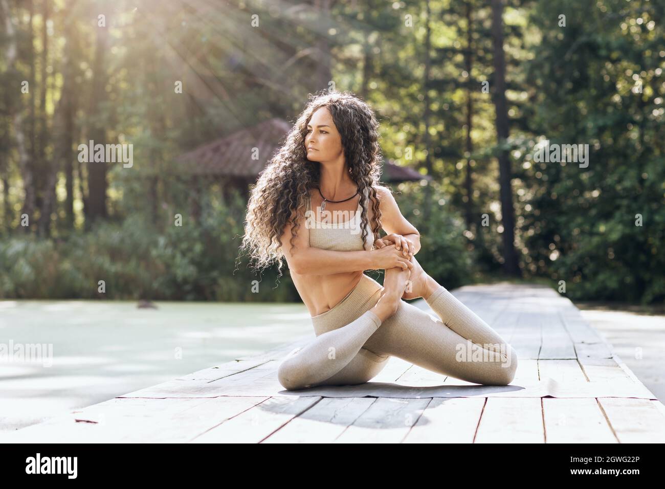 Attractive woman performs vamadevasana exercise, sage pose, sitting on a mat on a warm summer morning on a wooden bridge in the park Stock Photo