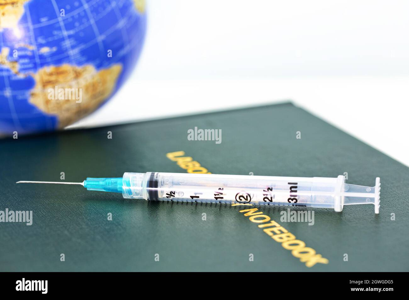 Injection needle placed with Lab Notebook and globe reflect worldwide nature of public health, medicine, pandemics, and healthcare industry Stock Photo