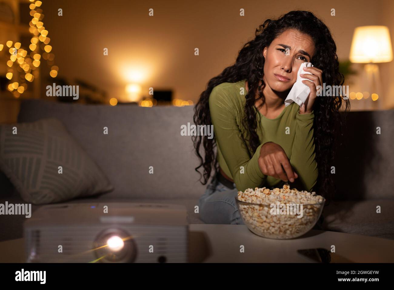Woman Crying Watching Sad Movie On Domestic Cinema Projector And Wiping Tears Sitting On Sofa At Home At Night. Female Depression And Loneliness, Ment Stock Photo