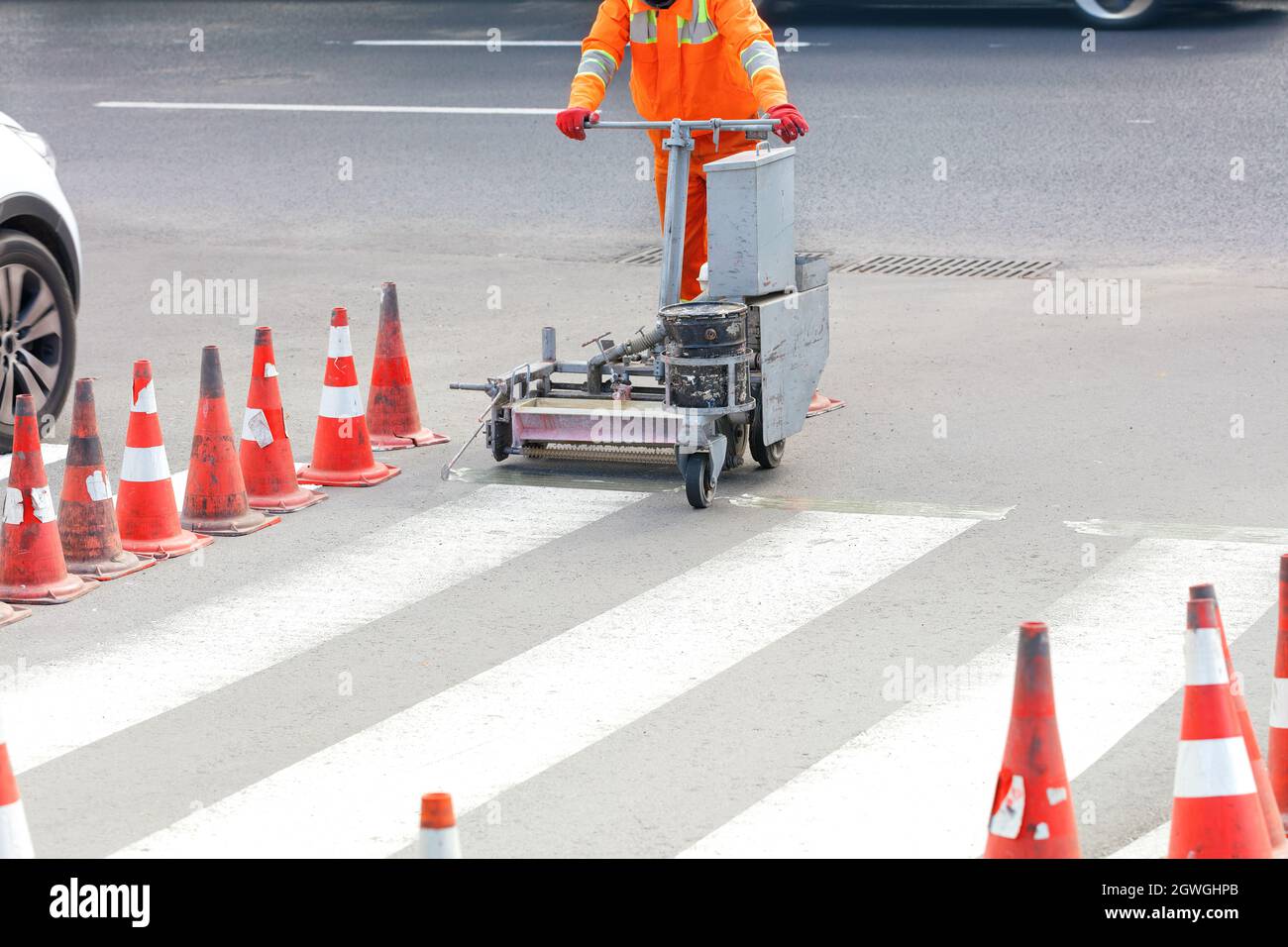 A road worker in orange overalls marks and renews pedestrian crossing lane markings on the road. Stock Photo
