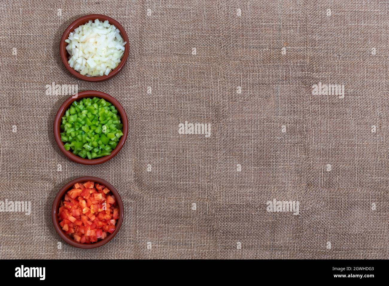 Background of rough fabric and brown color, with three small clay dishes with the ingredients of the sauce, onion, pepper and tomato.Copy space. Stock Photo