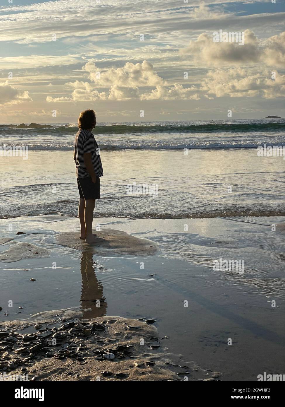Full length back view of a mature man standing on the beach late afternoon as the sun sets. Looking towards the horizon wearing shorts and t-shirt. Stock Photo
