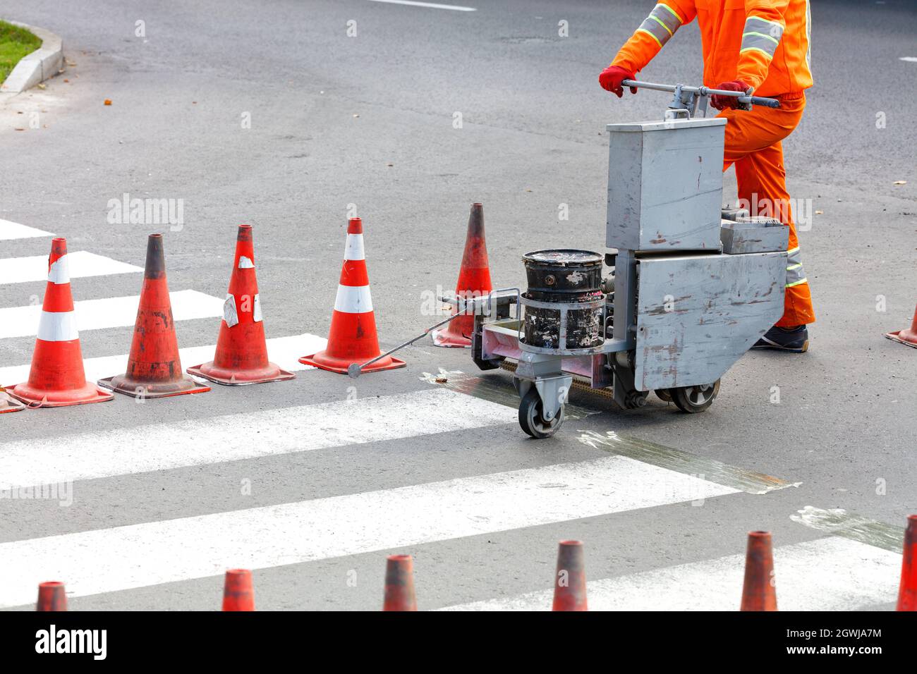 An orange overalls road worker renews pedestrian crosswalks on a road using a power trolley and traffic cones. Copy space. Stock Photo
