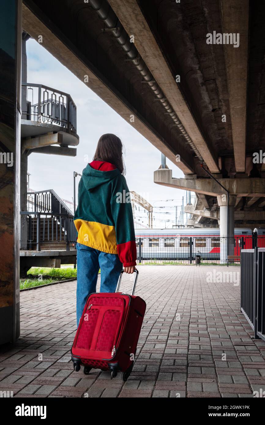 Rear view of a girl in bright clothes with a suitcase going to the train at the train station. A girl leaving by train on vacation, on a trip, on vaca Stock Photo