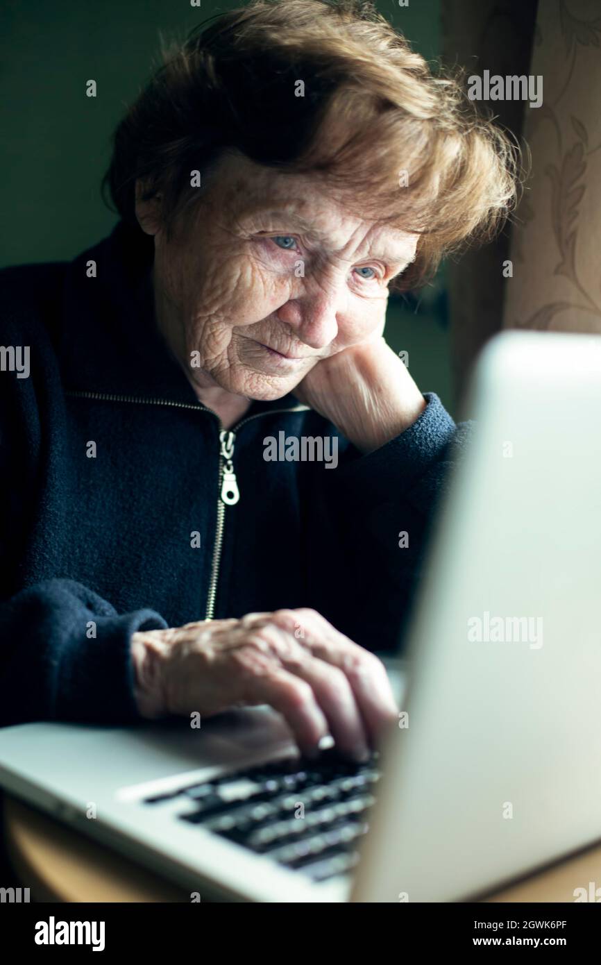 An old woman is typing on a laptop at her home. Stock Photo