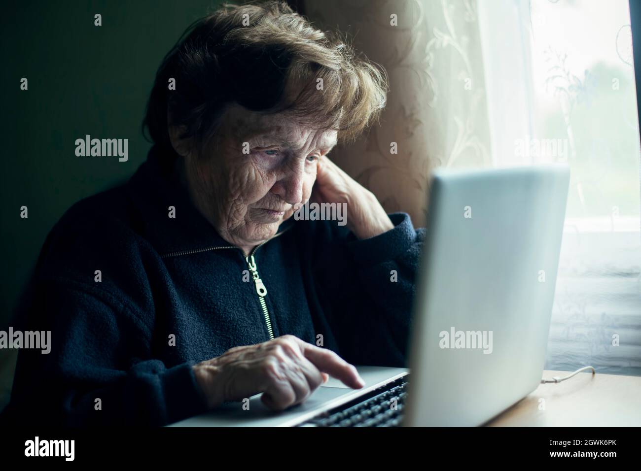 An old woman is typing on a laptop in her home. Stock Photo