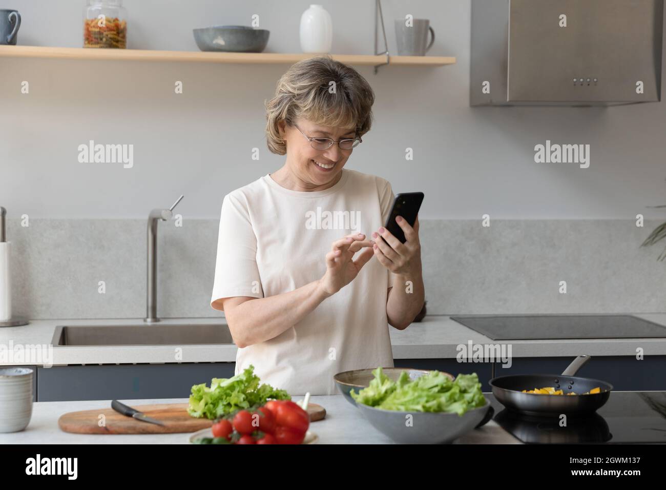 Happy middle aged woman using phone, cooking food. Stock Photo