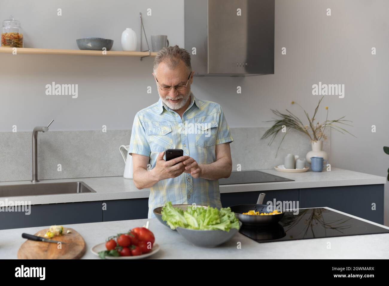 Joyful older mature man using cellphone, preparing food. Stock Photo