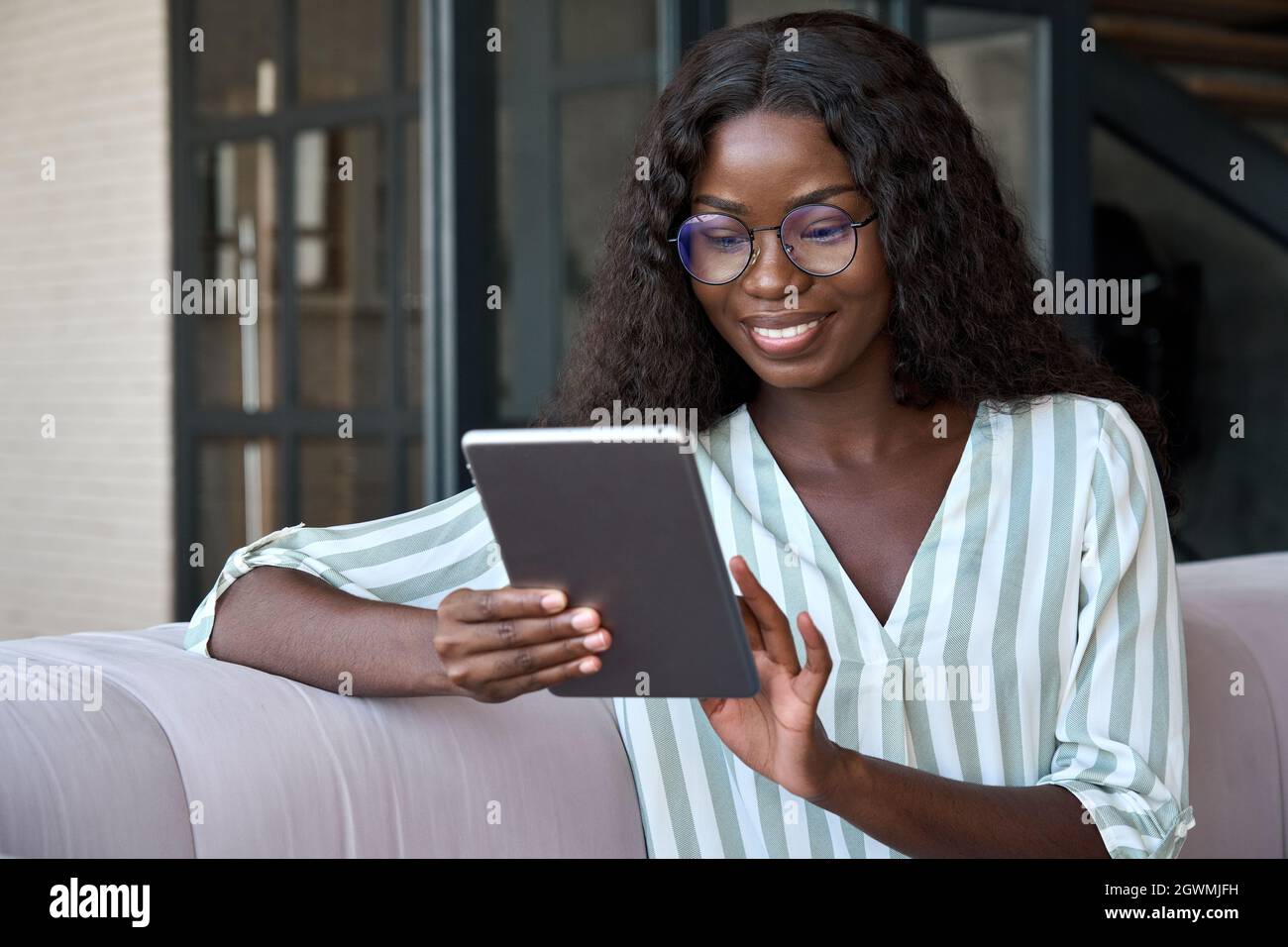 Afro American smiling young girl sitting on sofa at home holding tablet device. Stock Photo