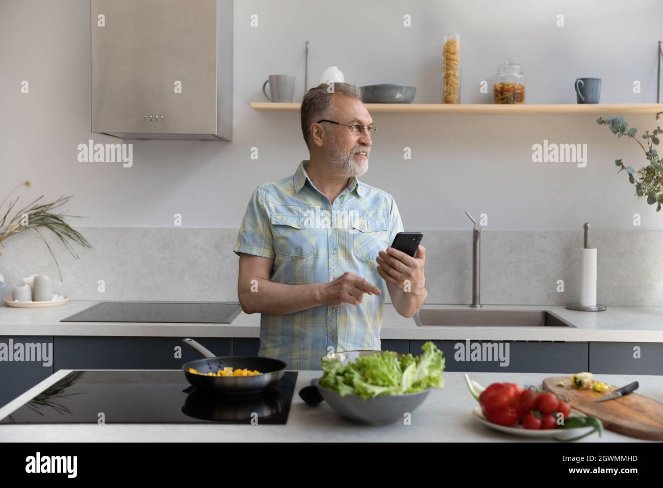 Smiling dreamy middle aged man using cellphone, cooking in kitchen. Stock Photo