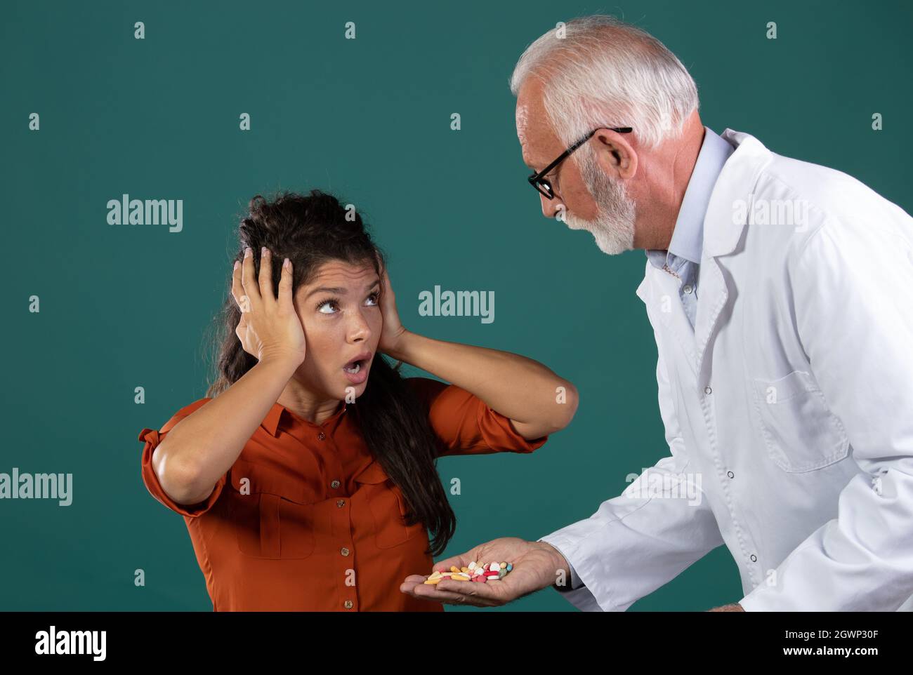 Senior experienced male doctor offering palm full of drugs to young patient that rejecting pills and holding hands on ears. Woman afraid of health car Stock Photo
