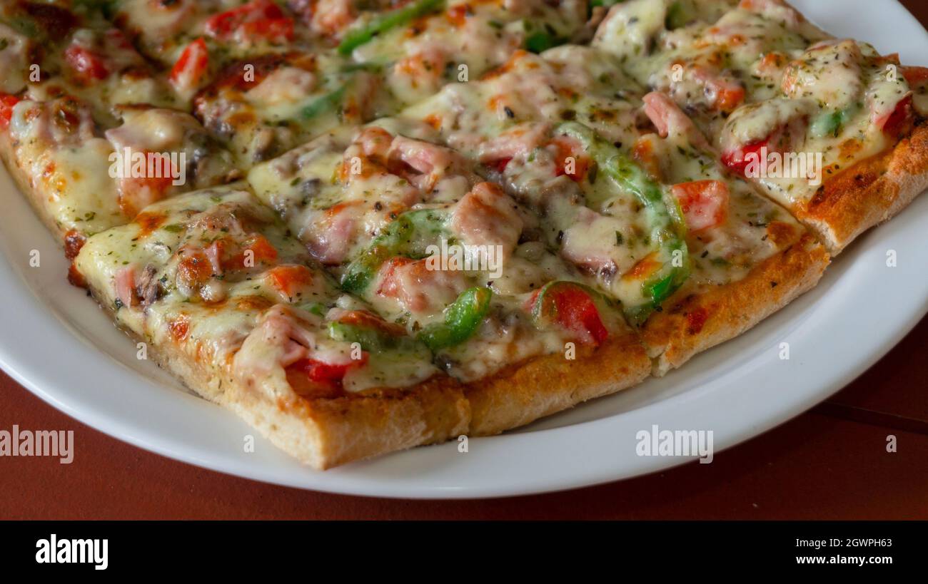 Close up on a white plate with a square pizza with ham, green pepper, tomato, olives and cheese cut into rectangular pieces on wooden table Stock Photo