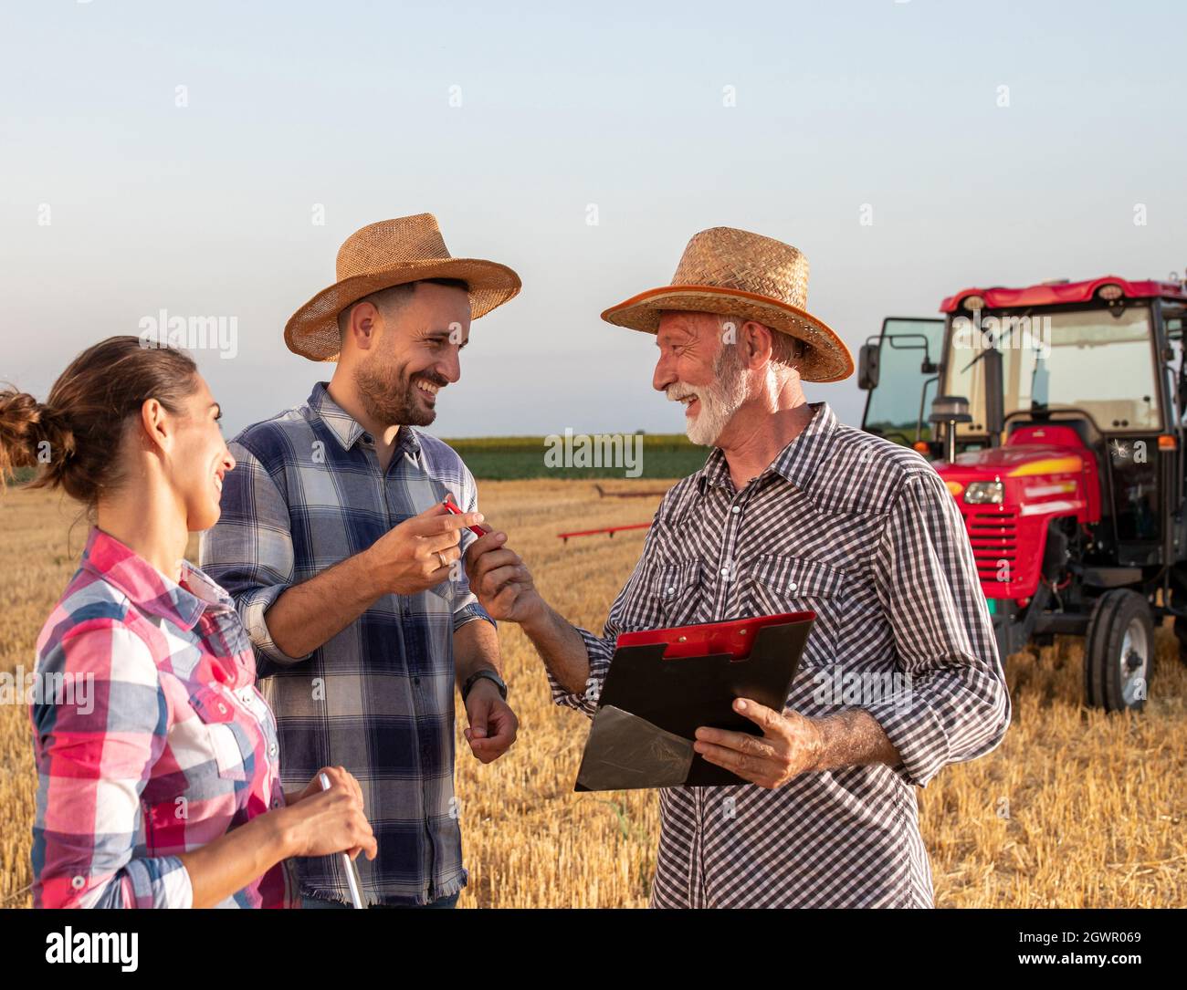 Elderly agronomist holding clipboard giving young farmer pen. Two farmers discussing business with insurance sales rep while standing in wheat field w Stock Photo