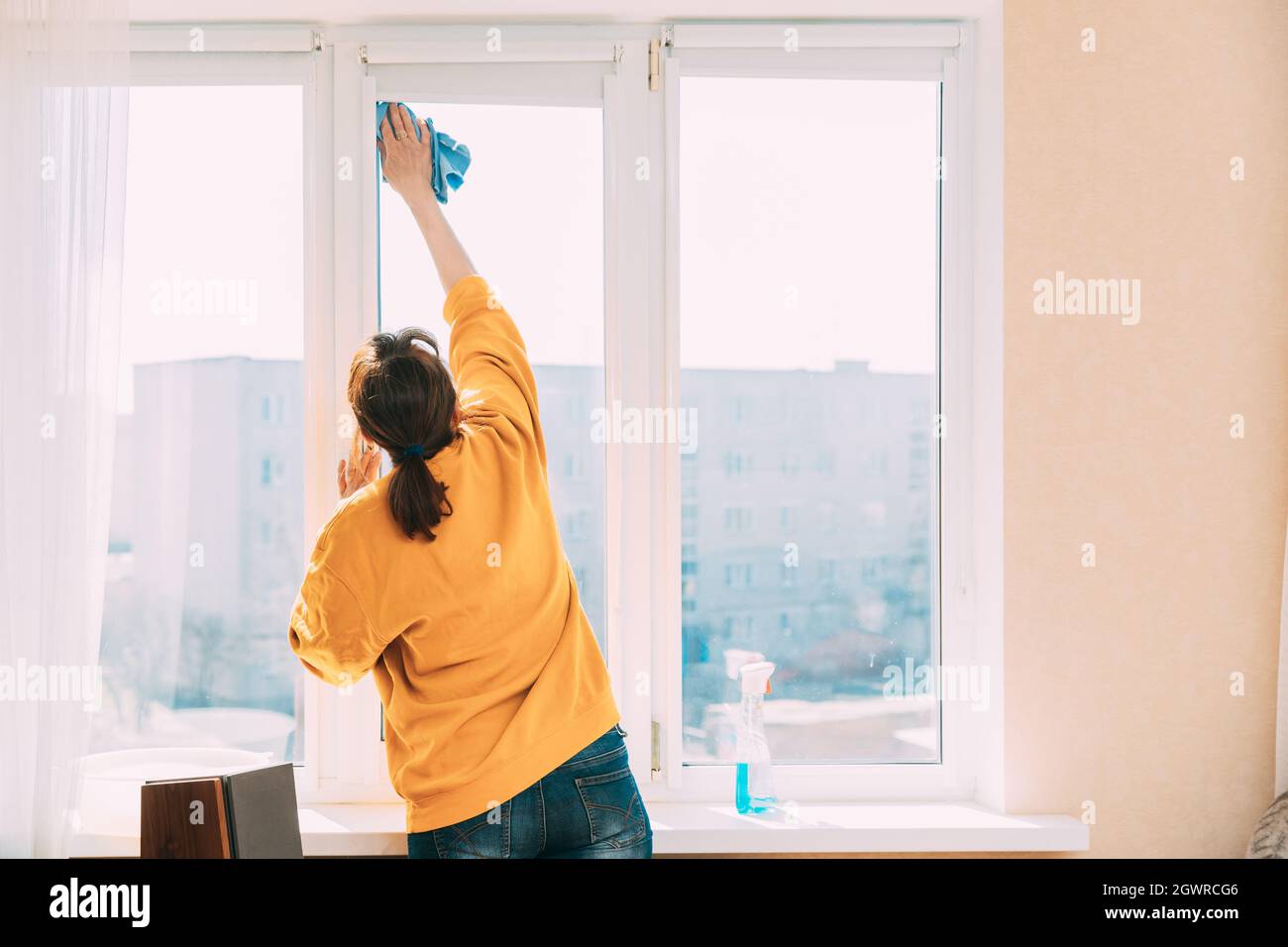 Woman Of Fifty In Yellow Sweater And Jeans Washes Dusty Window In Apartment. 50 Year Old Woman Cleans Windows From Stains Using Rag And Spray Cleaner Stock Photo