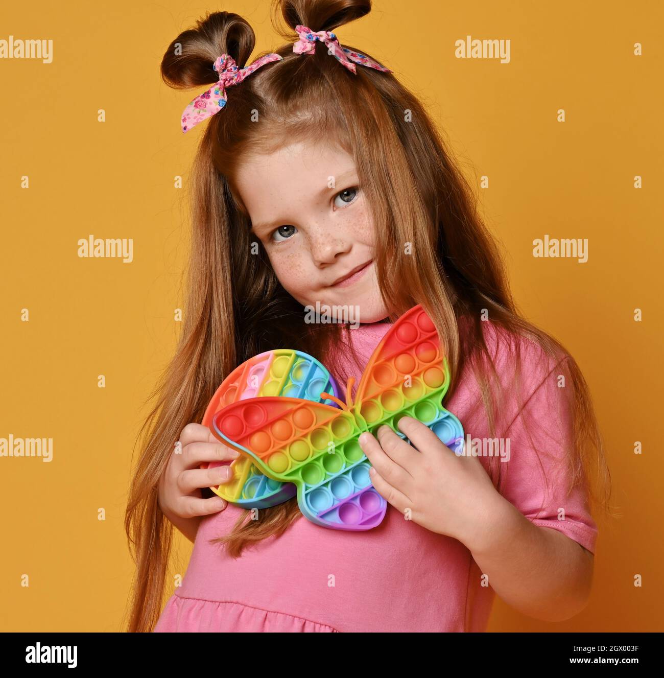 Red-haired kid girl in pink shirt stands hugging two new sensory rainbow color toys - butterfly shape and round pop it Stock Photo