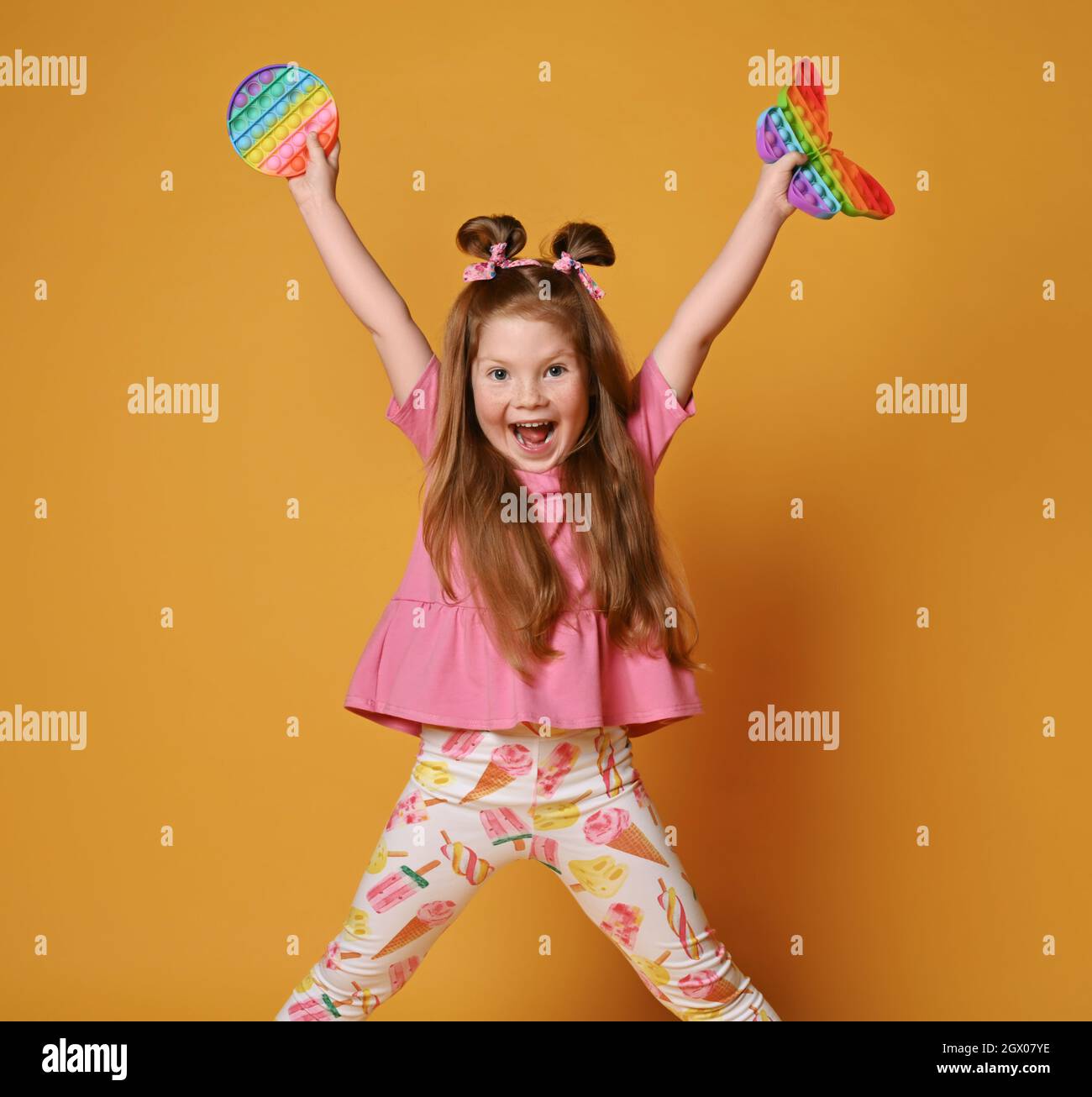 Kid girl in colorful clothes stands with legs wide apart, holds two sensory rainbow color toys - pop it up and screams Stock Photo