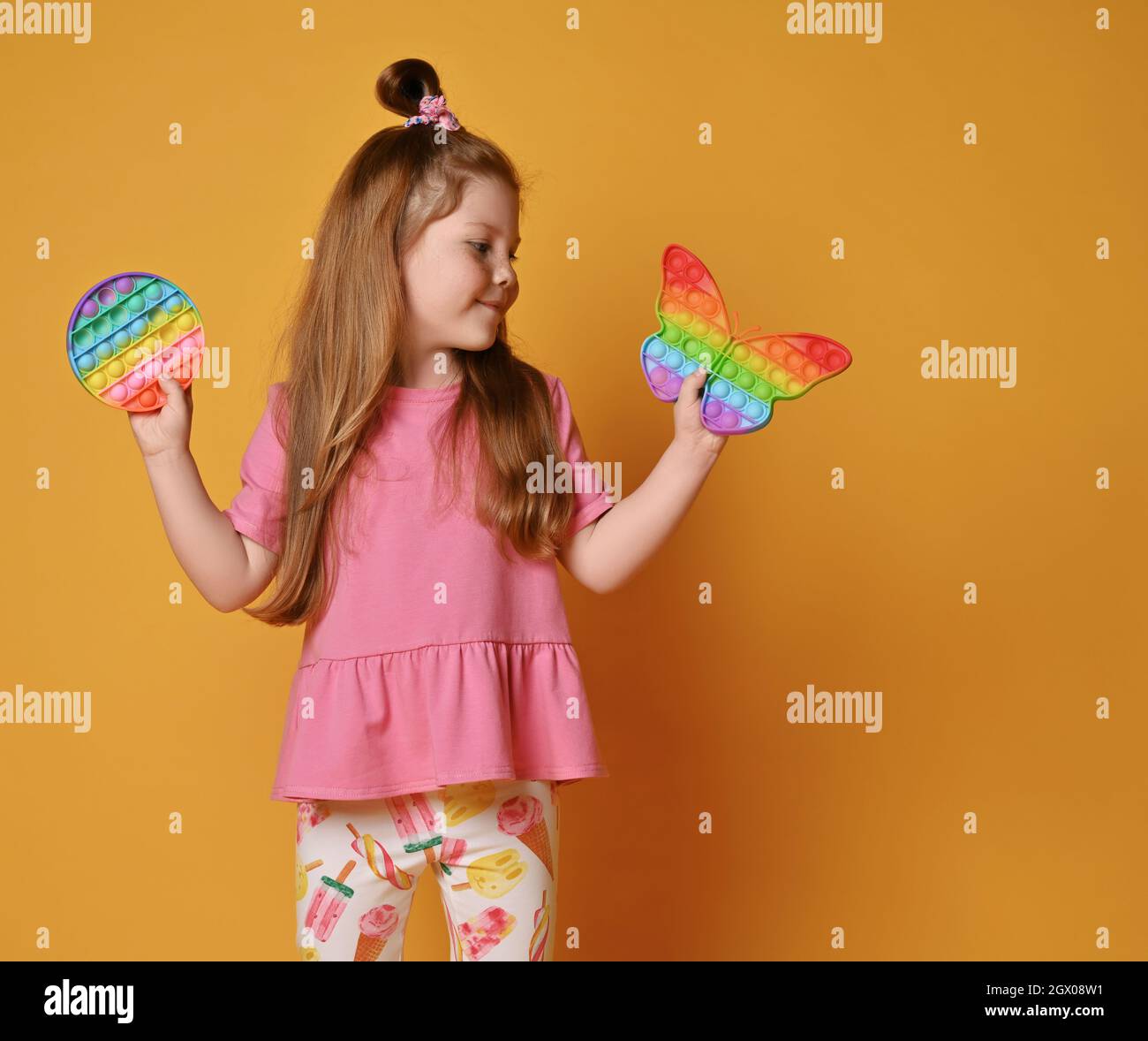 Kid girl in pink clothes shows two sensory rainbow color toys - pop it. Holds round and looks at butterfly shape one Stock Photo