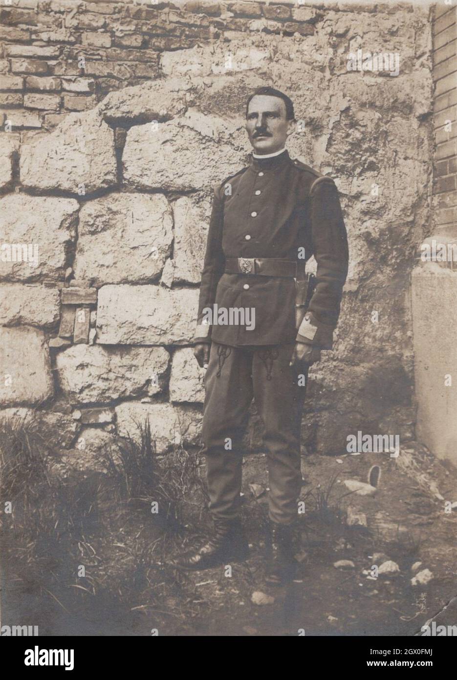 middle aged man posing for the camera in front of a castle at unknown place. This man is police officer or military officer but unrecogniseable that which empire's officer him. He has got black uniform, pistol holster, and a big massive impressive belt buckle. He hes got quite fashonable moustache. Period late 19th century early 20th century. Source: original photograph Stock Photo