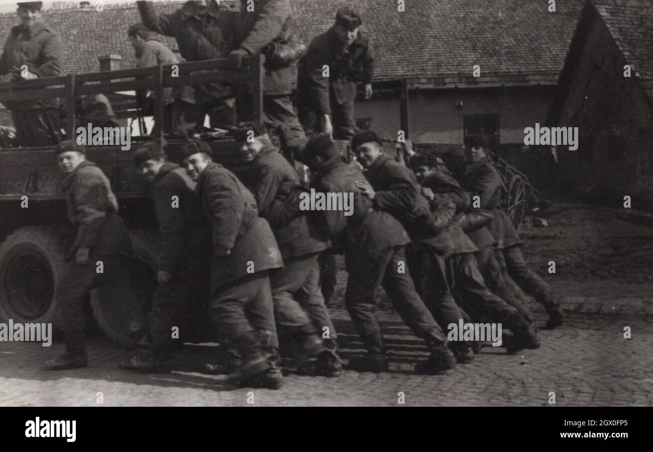 young army soldiers are pushing the military vehiche togedher which was breakdown.on the photograph. Possibly they just finished a practice at any shooting range. They are wearing uniforms and berets. Place: shooting range. Period: 1950's source: original photograph Stock Photo