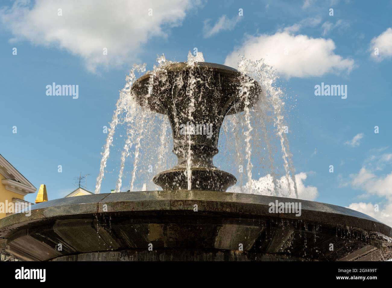 Water pours from a bowl of black marble fountain against a blue sky with small clouds. Stock Photo