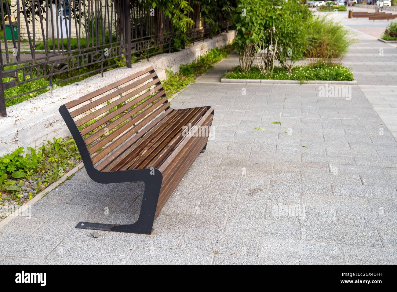 An interestingly designed street bench stands on the asphalt next to a metal fence. Stock Photo